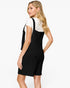A woman modeling a black knit overall-style dress with a white short-sleeve top layered underneath, shown from a three-quarters back view against a plain white studio background.