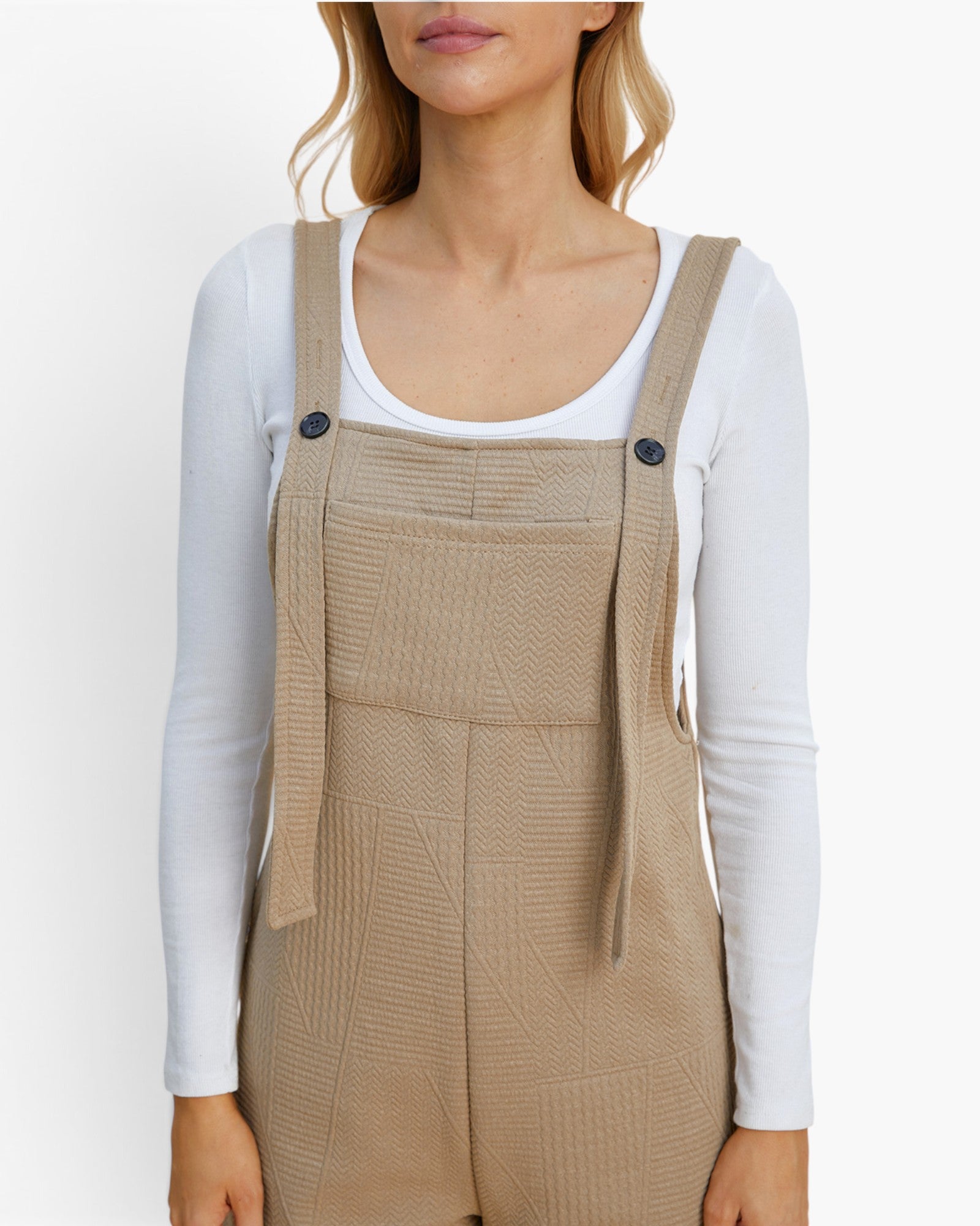 A woman wearing beige textured overall-style shorts over a white long-sleeve top, shown from the mouth to the upper thighs against a clean white background.