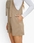 A woman wearing a beige ribbed knit sleeveless romper with a tie belt and side pockets, styled over a white long-sleeve top against a plain white background.