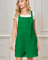 Woman wearing a green pinafore dress over a white shirt against a light gray wall.