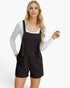 A woman modeling black textured overall shorts with adjustable shoulder straps over a white long-sleeve top, photographed against a clean white studio background.