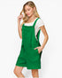 A woman modeling a bright green bib-style romper with shoulder straps, front pockets, and a white short-sleeve top underneath, photographed against a clean light gray studio background.