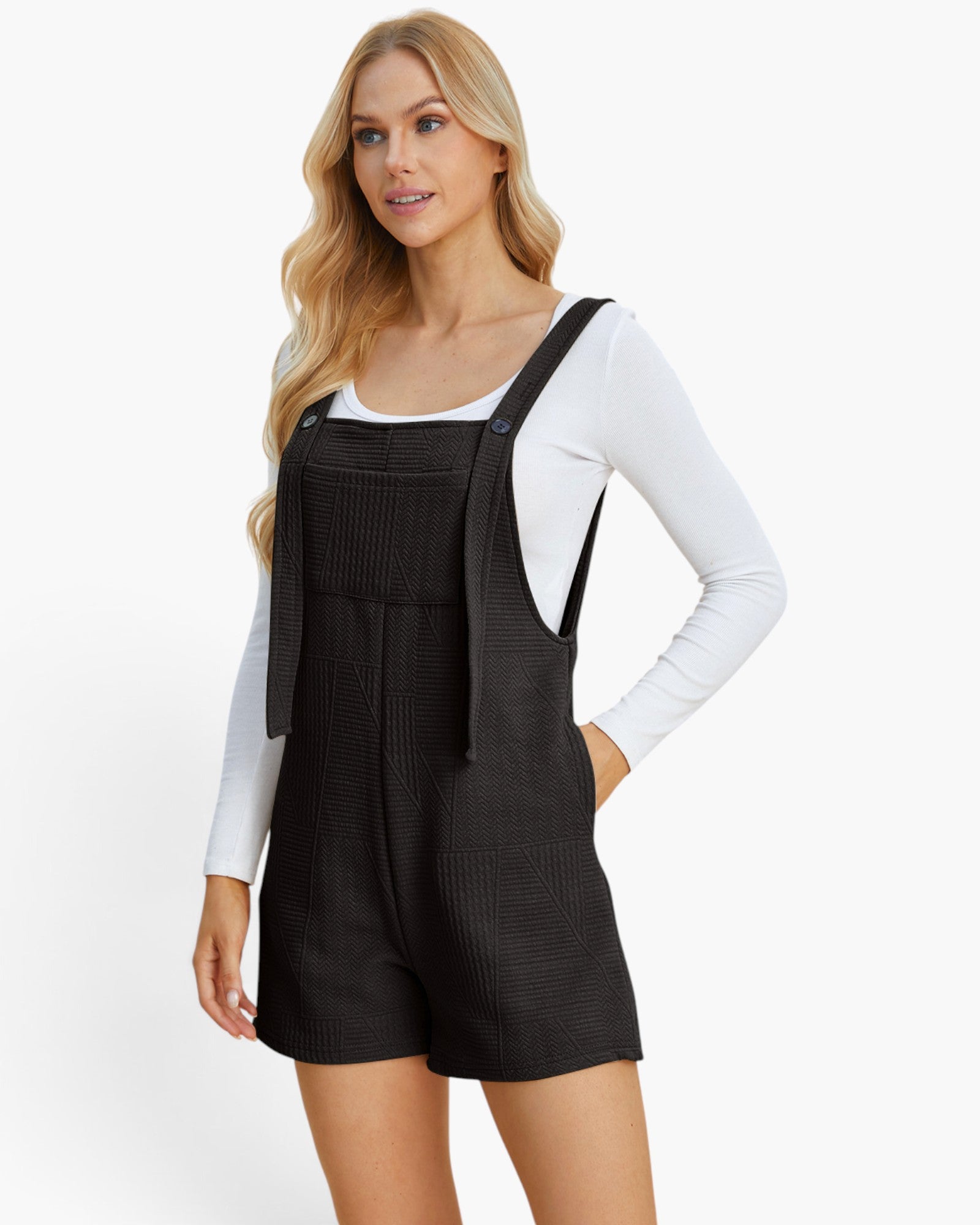 A blonde model wearing a black textured overall romper over a white long-sleeve top, photographed against a clean white studio background.