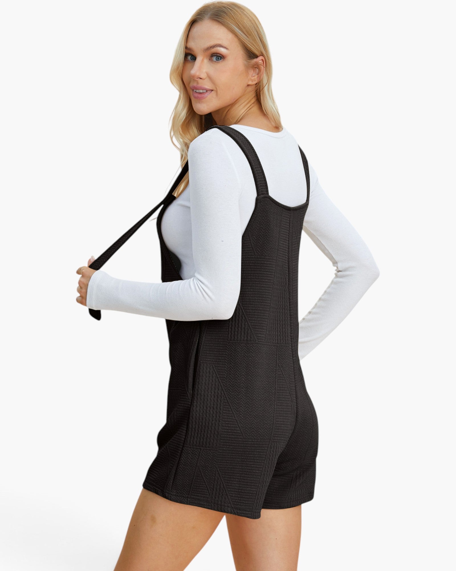A woman modeling a black ribbed overall romper with shoulder straps, worn over a white long-sleeve top, against a plain white studio background.