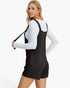 A woman modeling a black ribbed overall romper with shoulder straps, worn over a white long-sleeve top, against a plain white studio background.