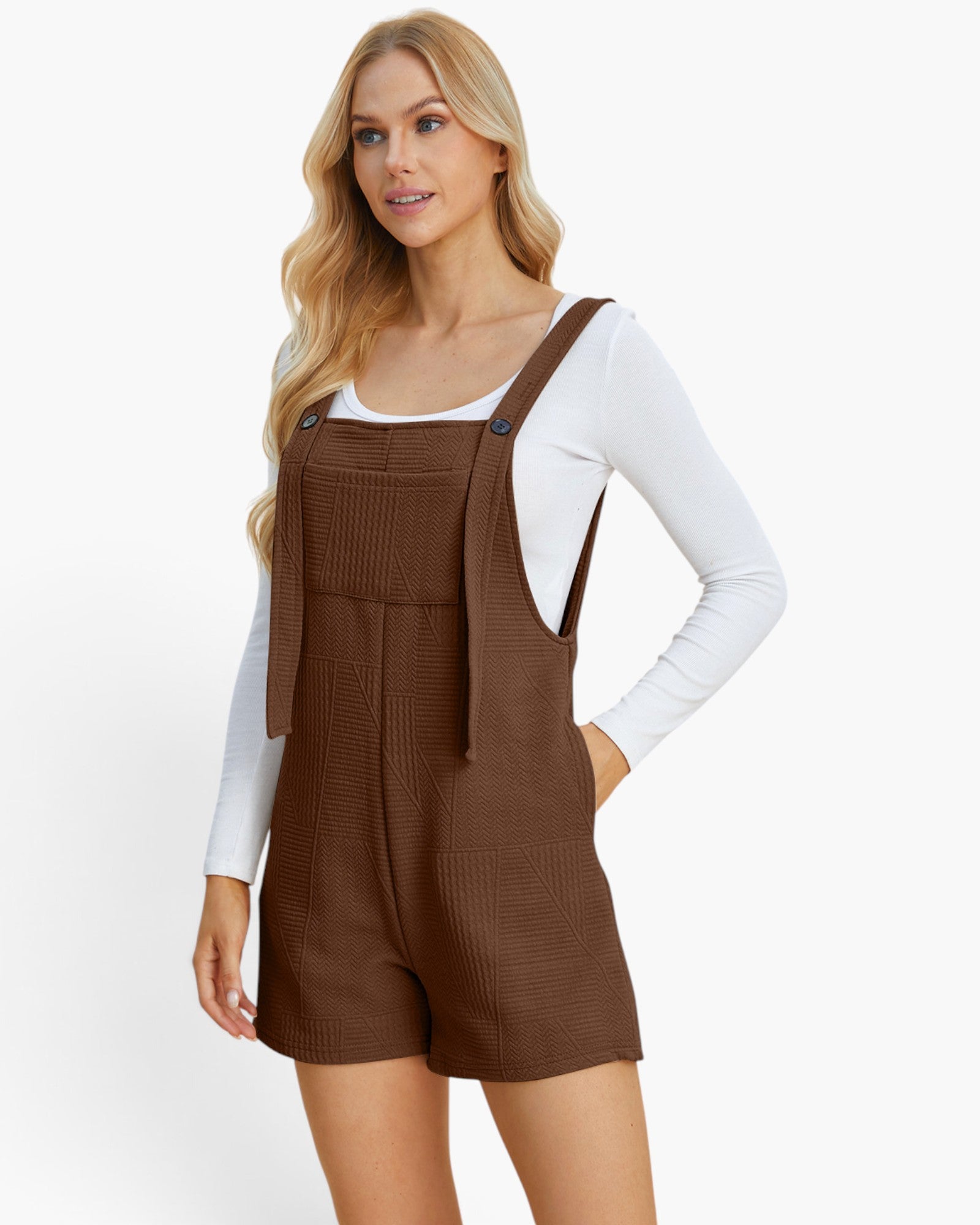 A blonde model wearing a brown textured overall romper with a white long-sleeve shirt underneath, photographed against a clean white studio background.