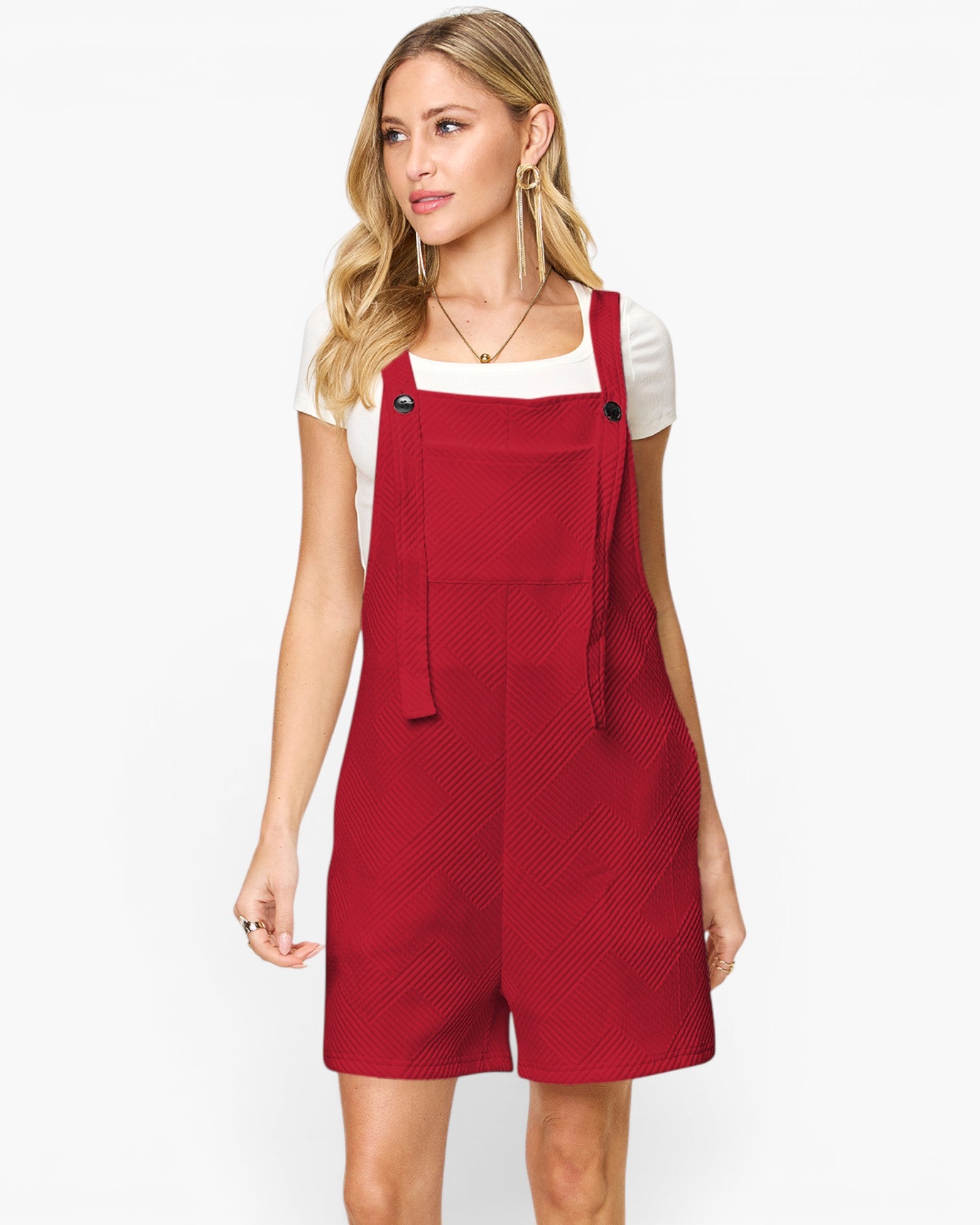 A woman modeling a red bib-style romper with shoulder straps and short legs, worn over a white short-sleeve top, against a plain light gray studio background.