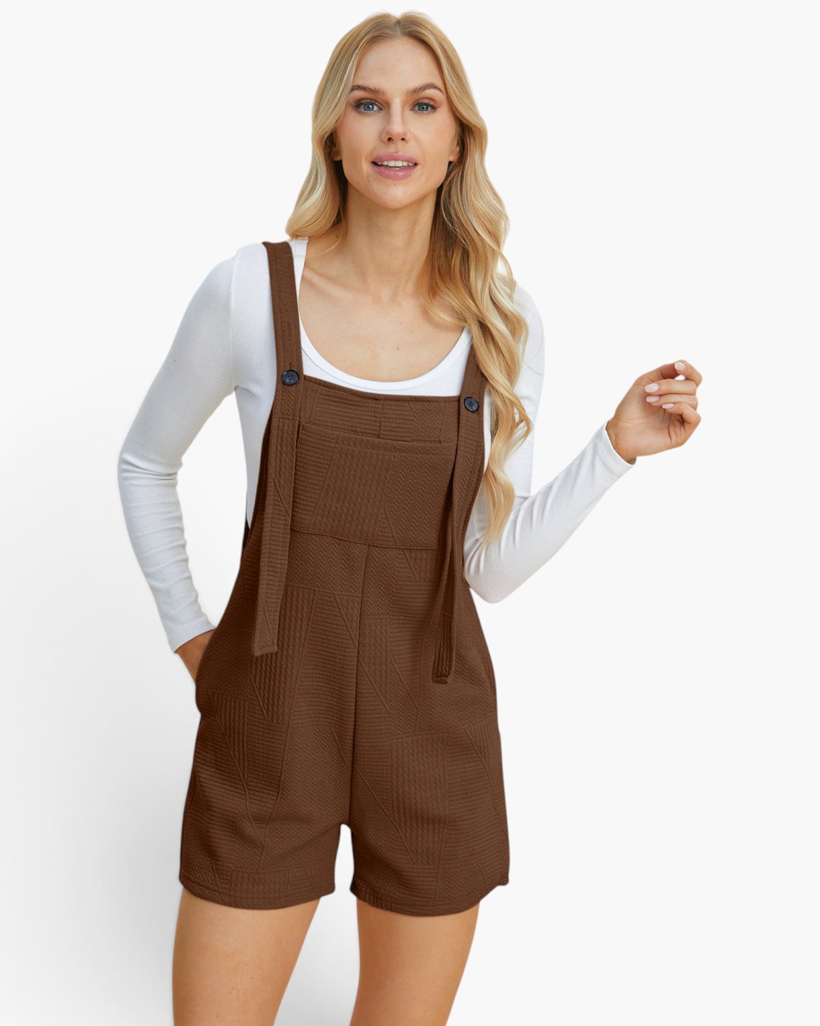A woman modeling a brown ribbed knit overall romper with shoulder straps and front pockets, worn over a white long-sleeve top, against a plain white studio background.