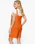A woman modeling a bright orange textured knit pinafore-style dress over a white short-sleeve top, shown from the back against a clean white studio background.