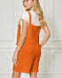Woman wearing an orange pinafore dress over a white top against a white paneled wall.