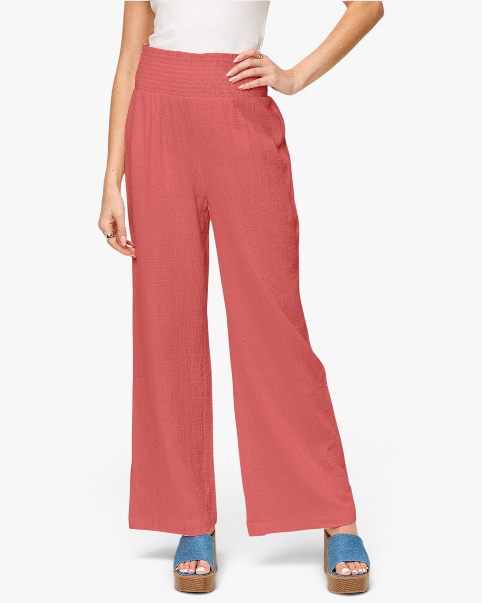 A model wearing high-waisted, wide-leg trousers in a dusty rose or terracotta pink color, paired with a white top and blue platform sandals, against a plain white studio background.