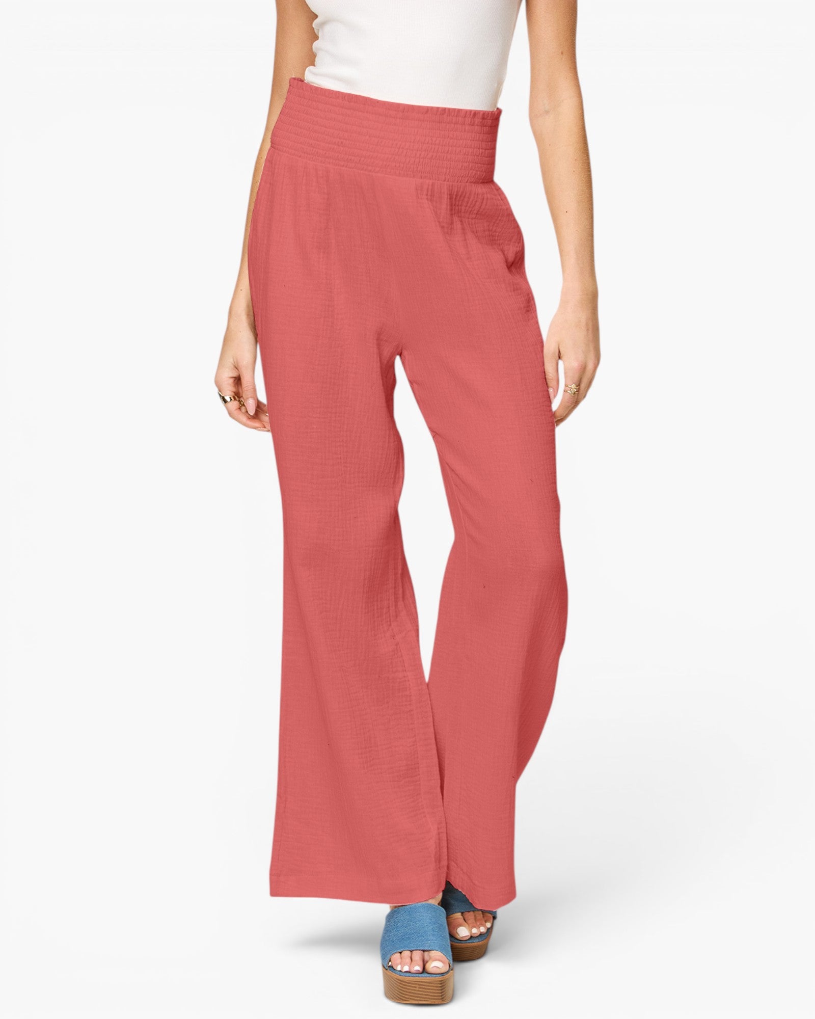 A model wearing a pair of wide-leg, high-waisted pants in a dusty rose or coral pink color, paired with a white top and blue platform sandals, against a plain white studio background.