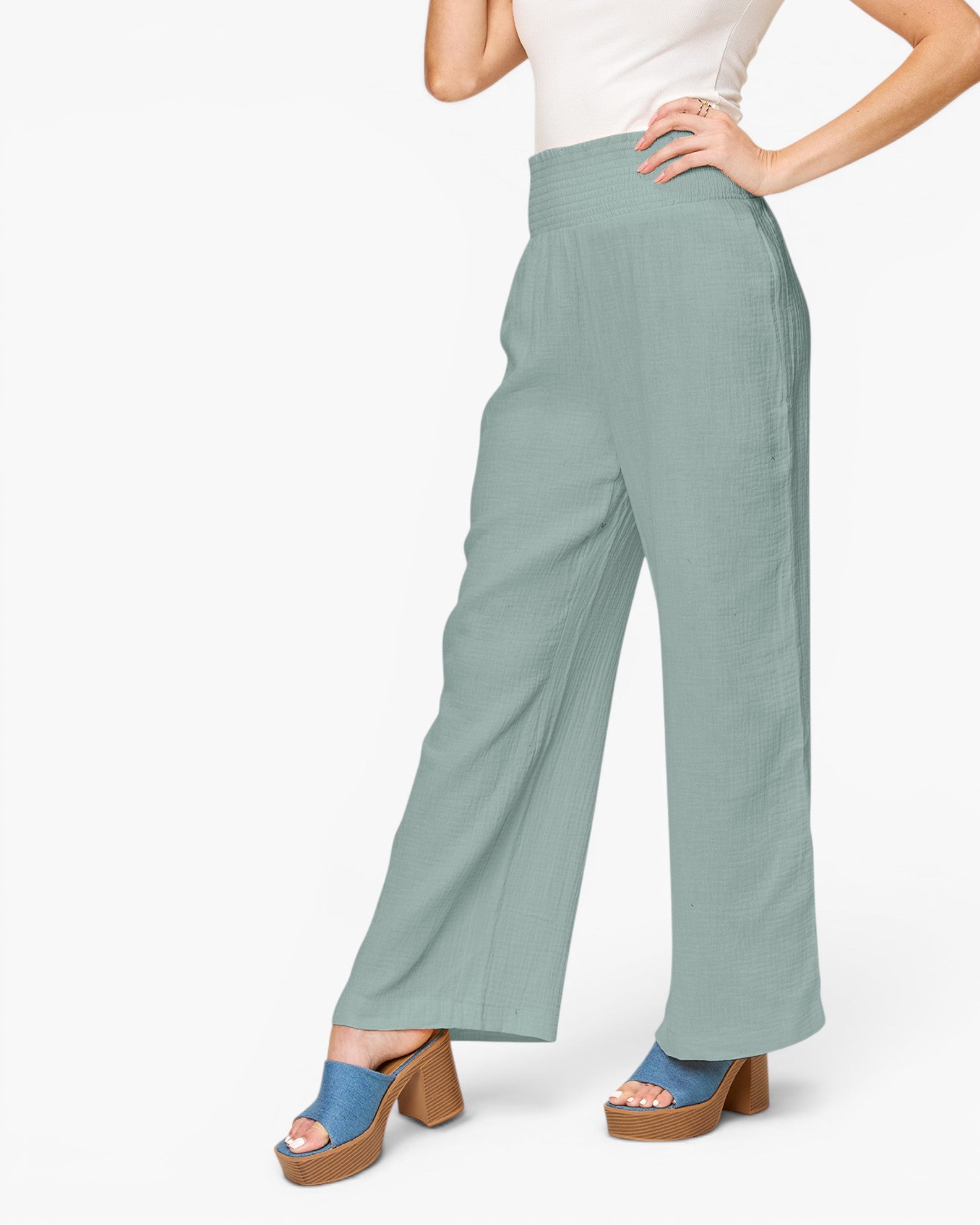A model wearing high-waisted, wide-leg pants in a dusty sage green color, styled with a white top and blue platform sandals against a plain white background.