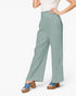 A model wearing high-waisted, wide-leg pants in a dusty sage green color, styled with a white top and blue platform sandals against a plain white background.