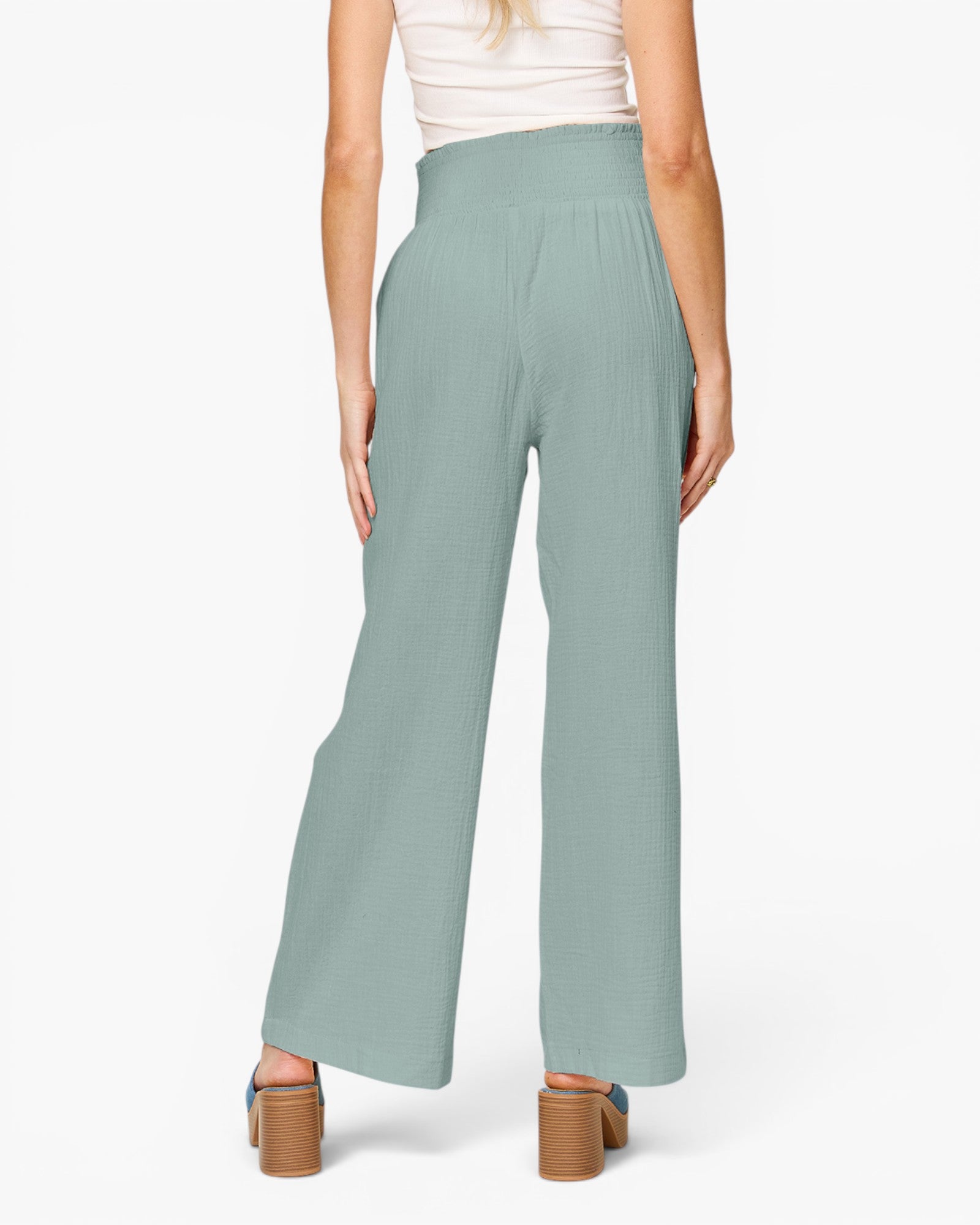 A model shown from the back wearing high-waisted, wide-leg pants in a dusty sage green color, paired with a white top and brown block-heel sandals, against a plain white studio background.