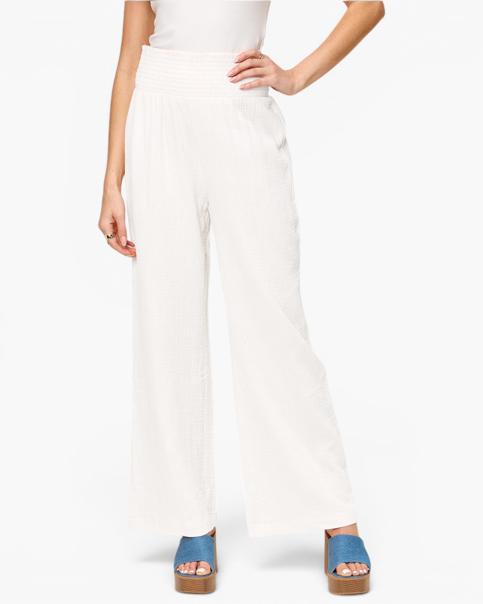 A model wearing a pair of high-waisted, wide-leg white pants with a textured, pleated fabric, styled with blue platform sandals against a plain white background.