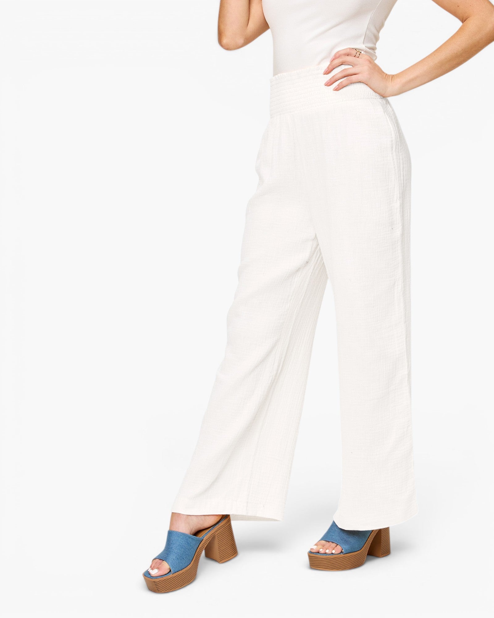 A model wearing high-waisted, wide-leg white pants with a white sleeveless top and blue platform mule sandals, shown from the waist down against a plain white background.