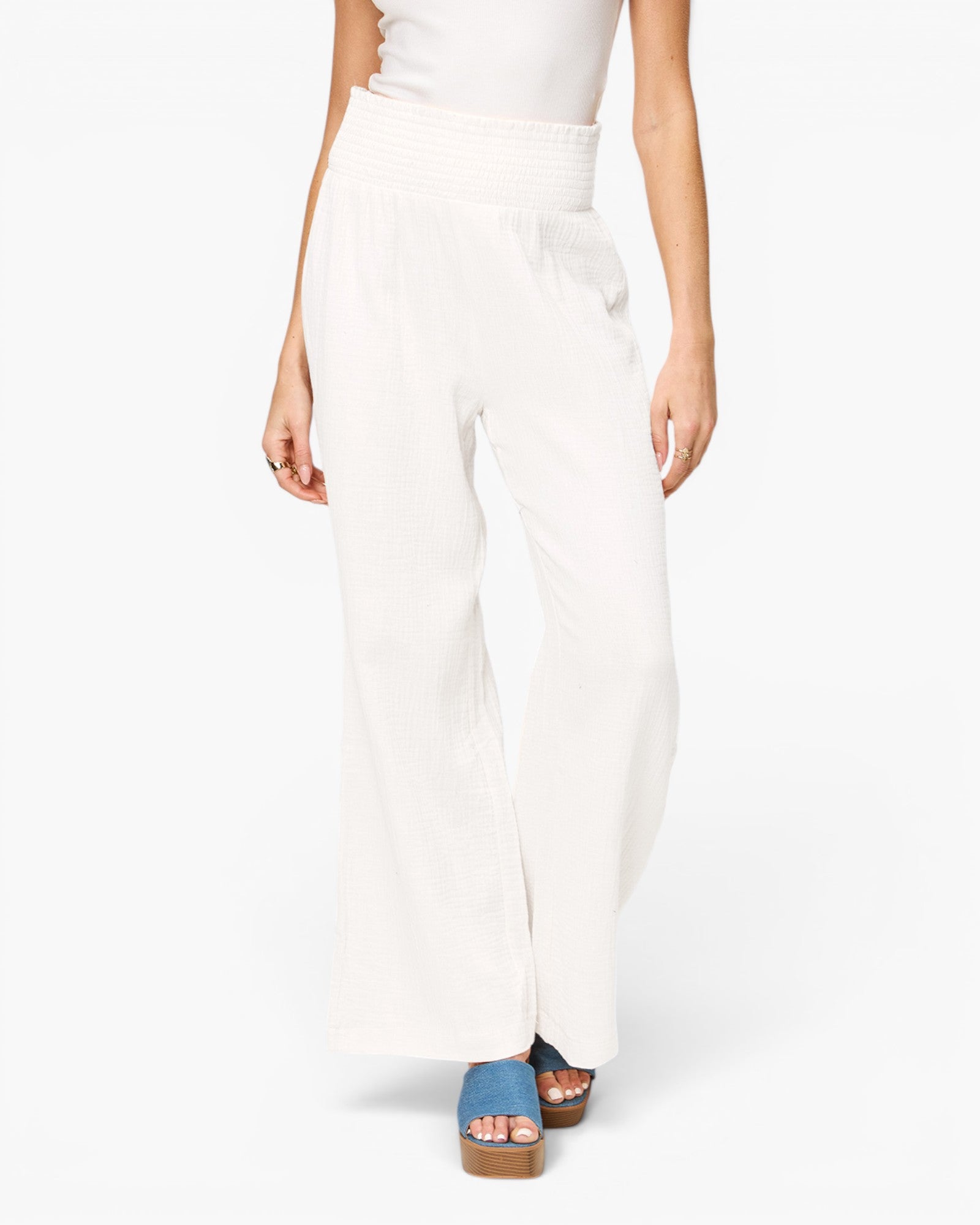 A model from the waist down wearing high-waisted, wide-leg white pants and blue platform sandals against a plain white studio background.