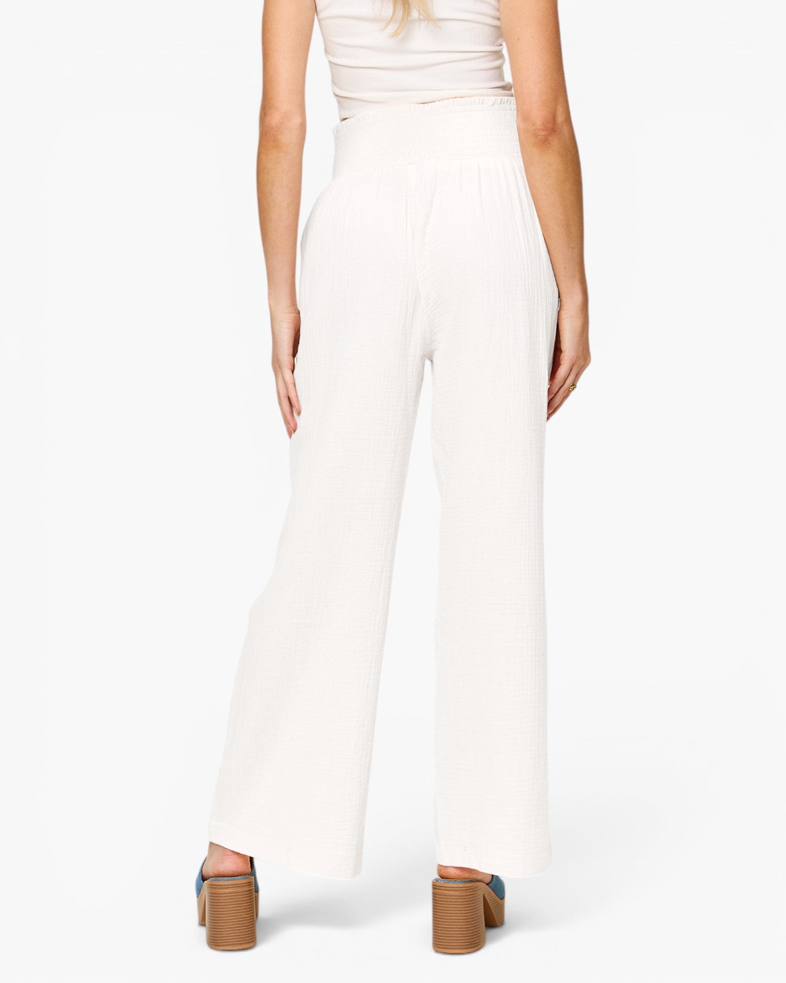 A model shown from the back wearing high-waisted, wide-leg white pants with a matching white top and brown block-heel sandals, set against a plain white studio background.