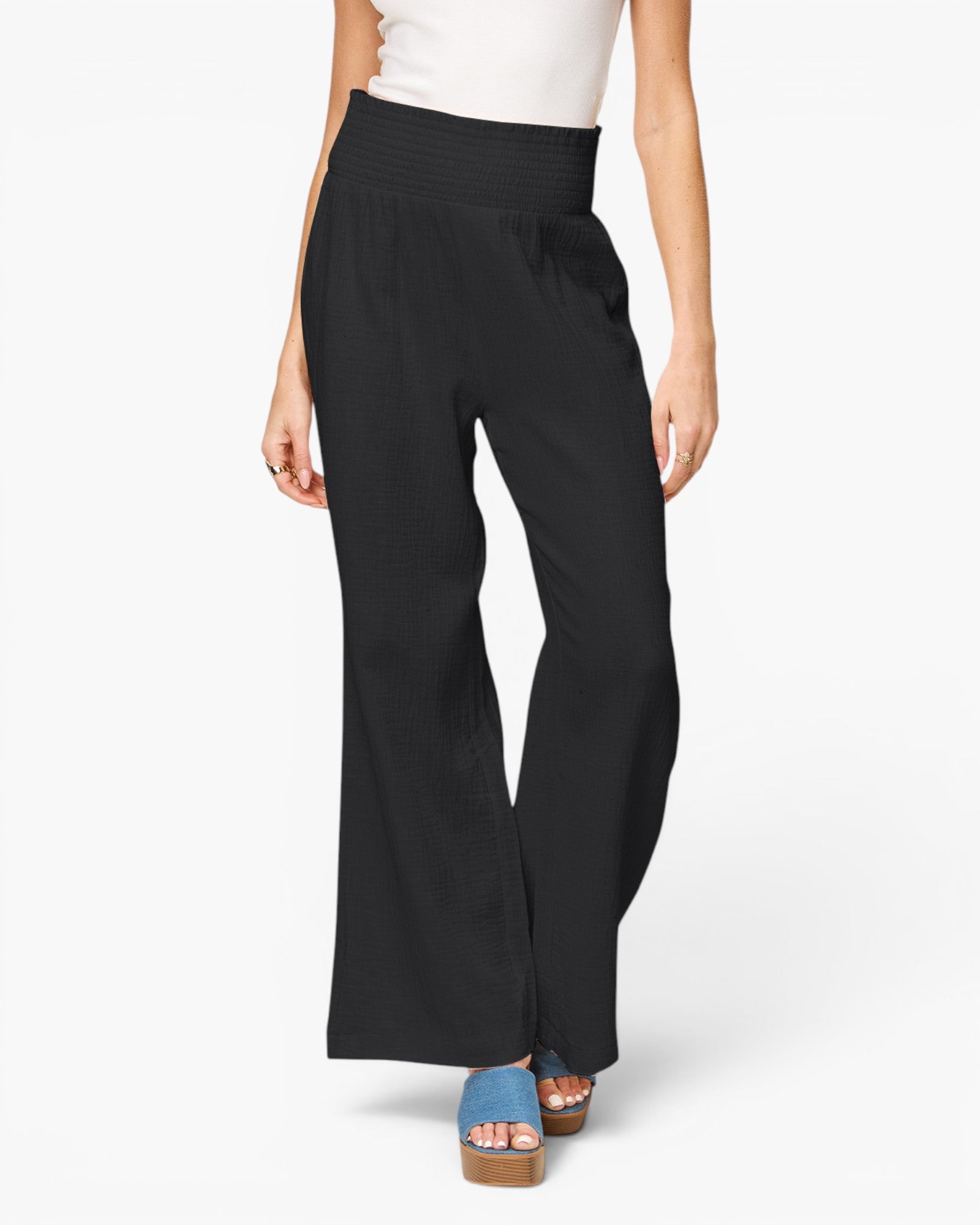 A model wearing black wide-leg cropped pants with a smocked elastic waistband, paired with a white top and blue platform sandals, shown from the waist down on a light gray background.