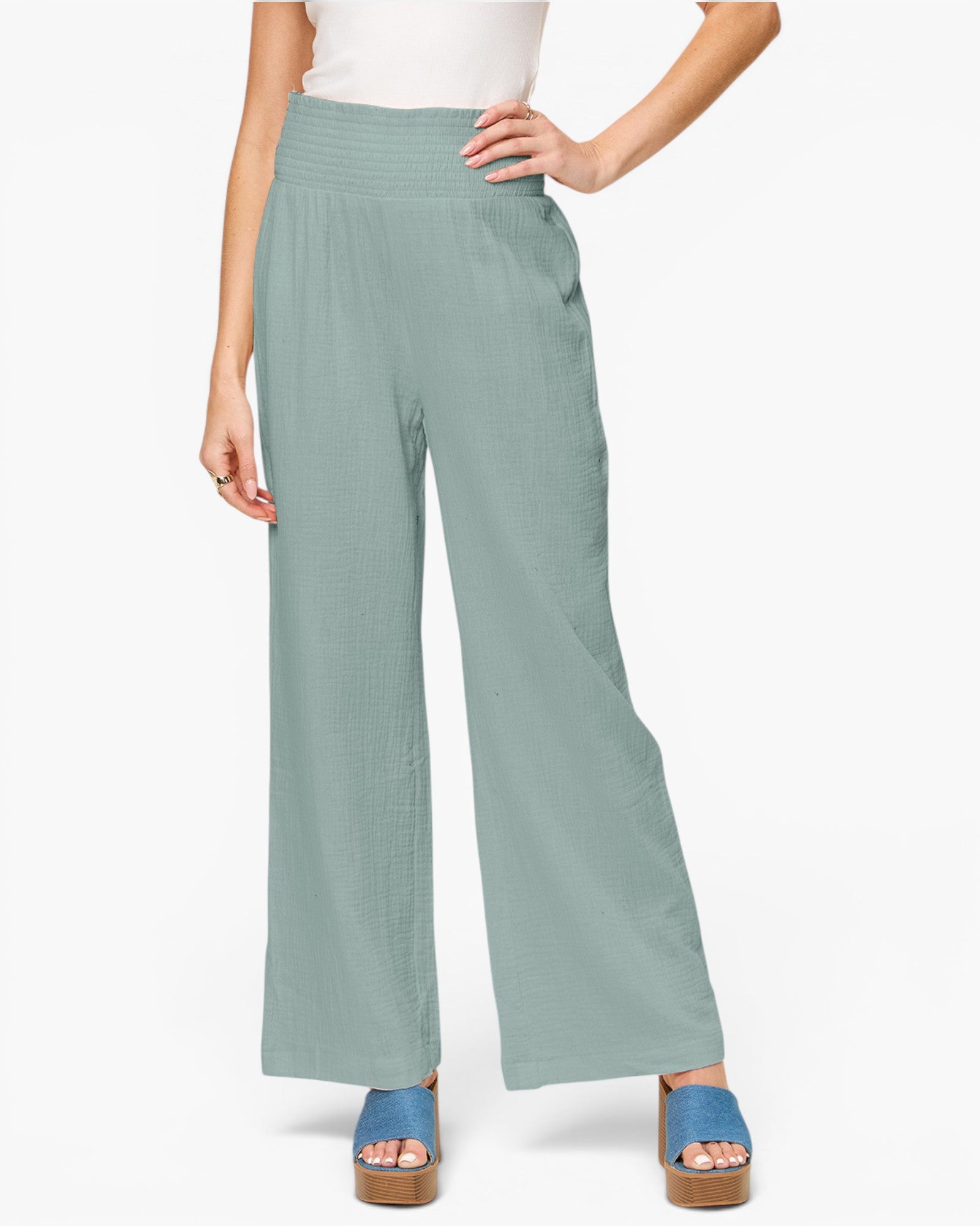 A model wearing high-waisted, wide-leg pants in a dusty sage green color, paired with a white top and blue platform sandals, against a plain white background.