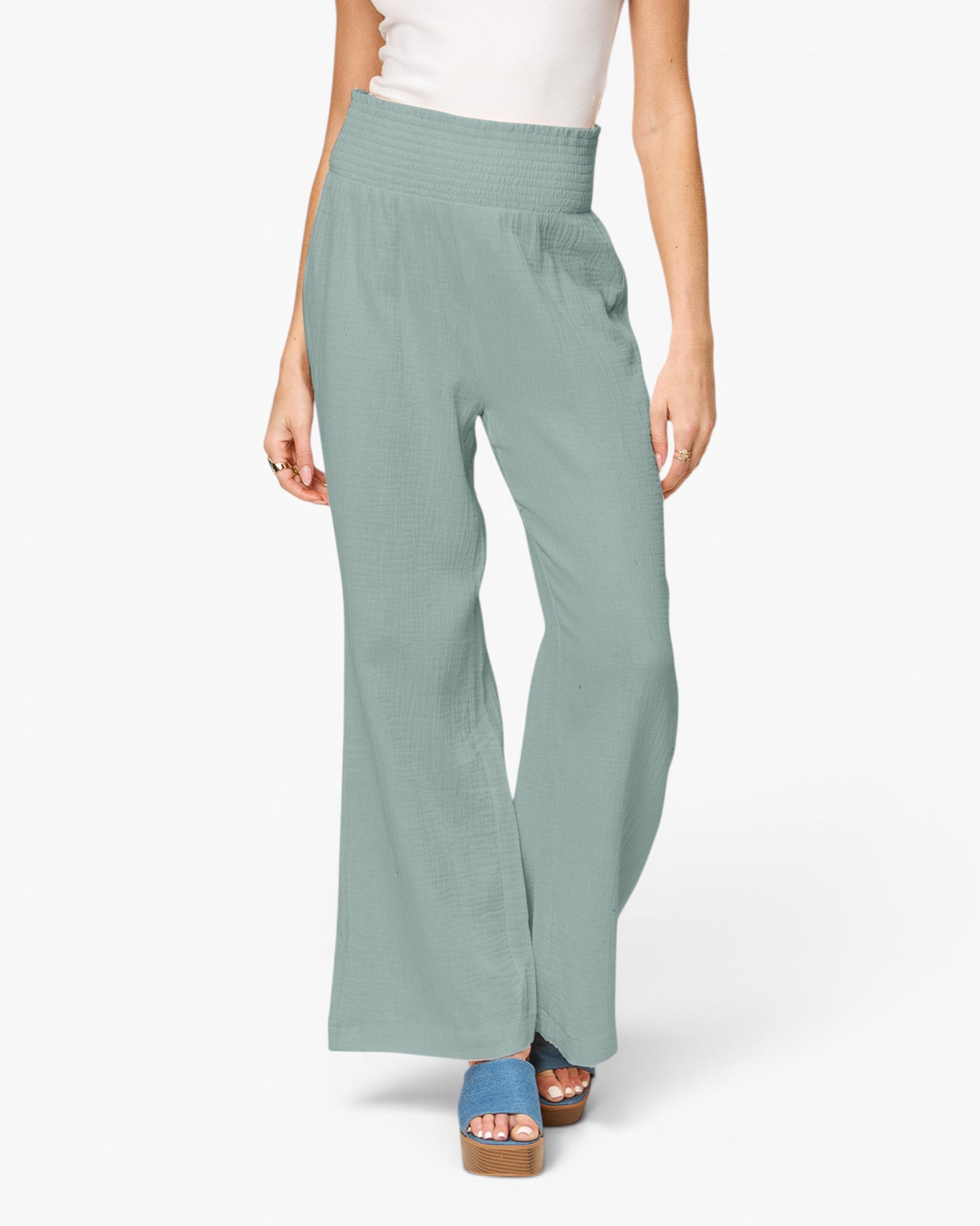 A model from the waist down wearing high-waisted, wide-leg pants in a dusty sage green color, paired with a white top and blue platform sandals, against a plain white studio background.