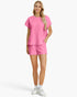 A woman modeling a matching bright pink two-piece lounge set with a short-sleeve top and shorts, featuring a textured waffle pattern, against a light gray studio background.