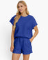 A woman modeling a matching royal blue two-piece lounge set featuring a short-sleeve top and shorts with a textured, quilted pattern, set against a plain white background.