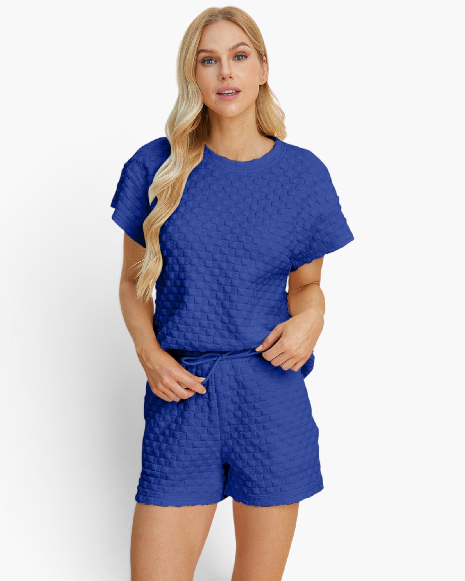 A blonde woman modeling a matching royal blue two-piece set consisting of a short-sleeve top and shorts, both featuring a textured, quilted waffle pattern. She is standing against a clean, light off-white studio background.