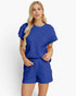 A blonde woman modeling a matching royal blue two-piece set consisting of a short-sleeve top and shorts, both featuring a textured, quilted waffle pattern. She is standing against a clean, light off-white studio background.