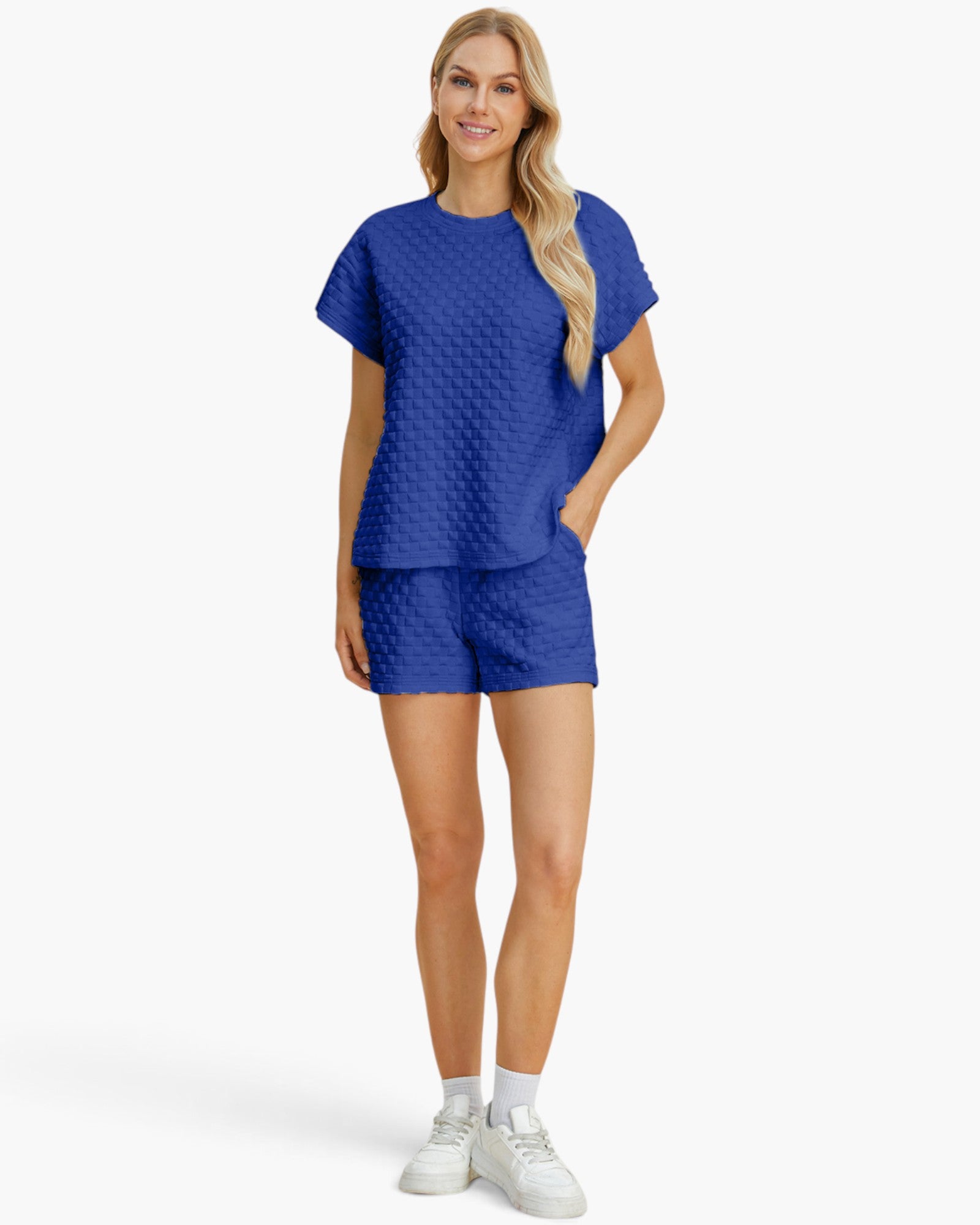 A woman modeling a matching royal blue two-piece lounge set featuring a short-sleeve top and shorts with a textured geometric pattern, paired with white sneakers against a light gray background.