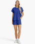 A woman modeling a matching royal blue two-piece lounge set featuring a short-sleeve top and shorts with a textured geometric pattern, paired with white sneakers against a light gray background.