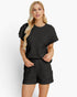 A blonde woman modeling a black short-sleeve romper with a textured, quilted pattern against a plain white studio background.