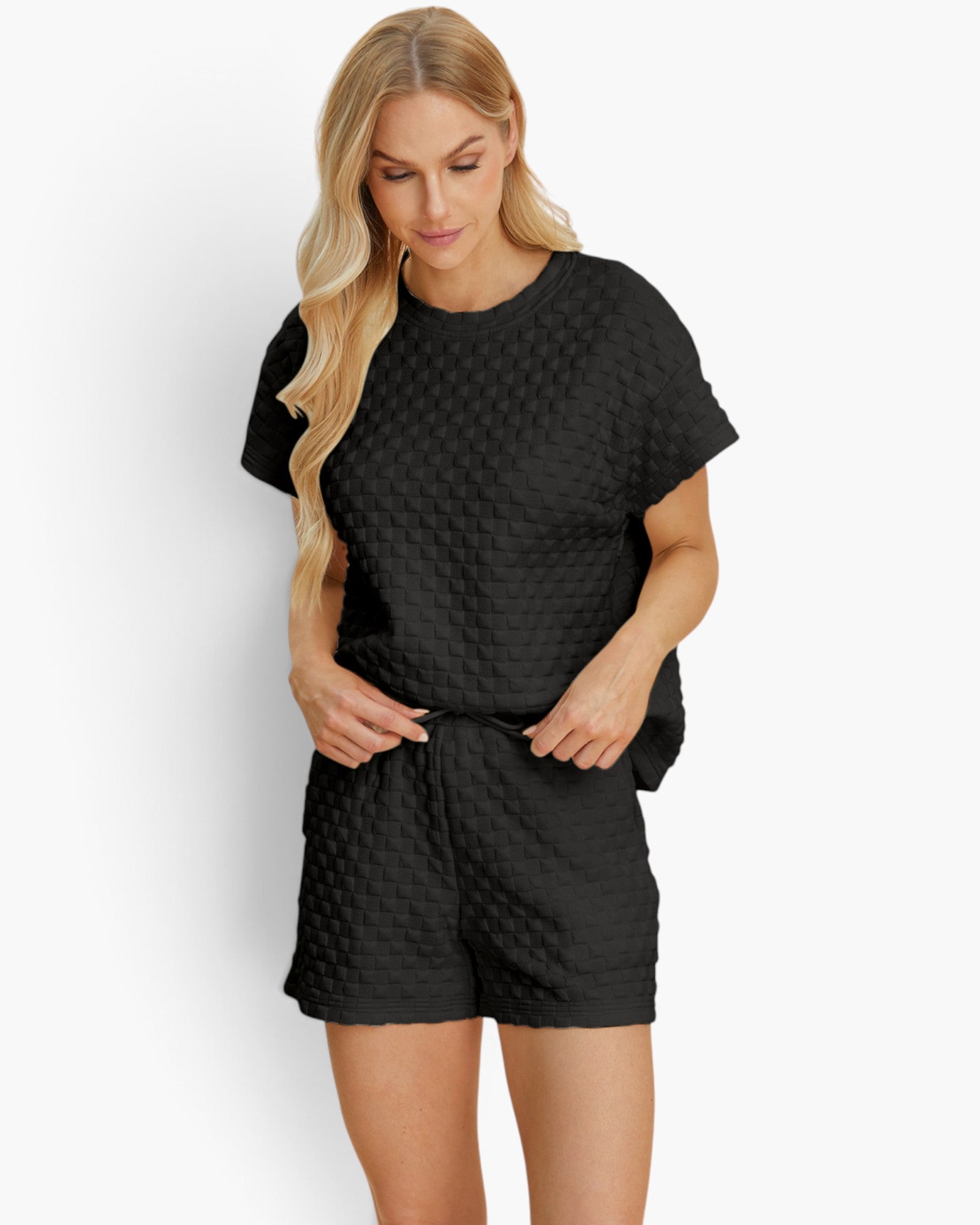 A woman modeling a black two-piece lounge set featuring a short-sleeve top and matching shorts, both with a textured waffle-weave pattern, against a plain white background.