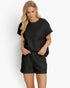 A woman modeling a black two-piece lounge set featuring a short-sleeve top and matching shorts, both with a textured waffle-weave pattern, against a plain white background.