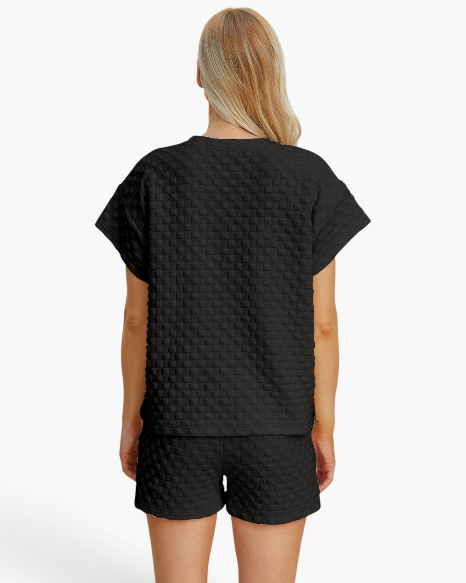A woman viewed from the back wearing a black short-sleeve top and matching black shorts with a subtle embossed geometric pattern, against a plain light gray studio background.