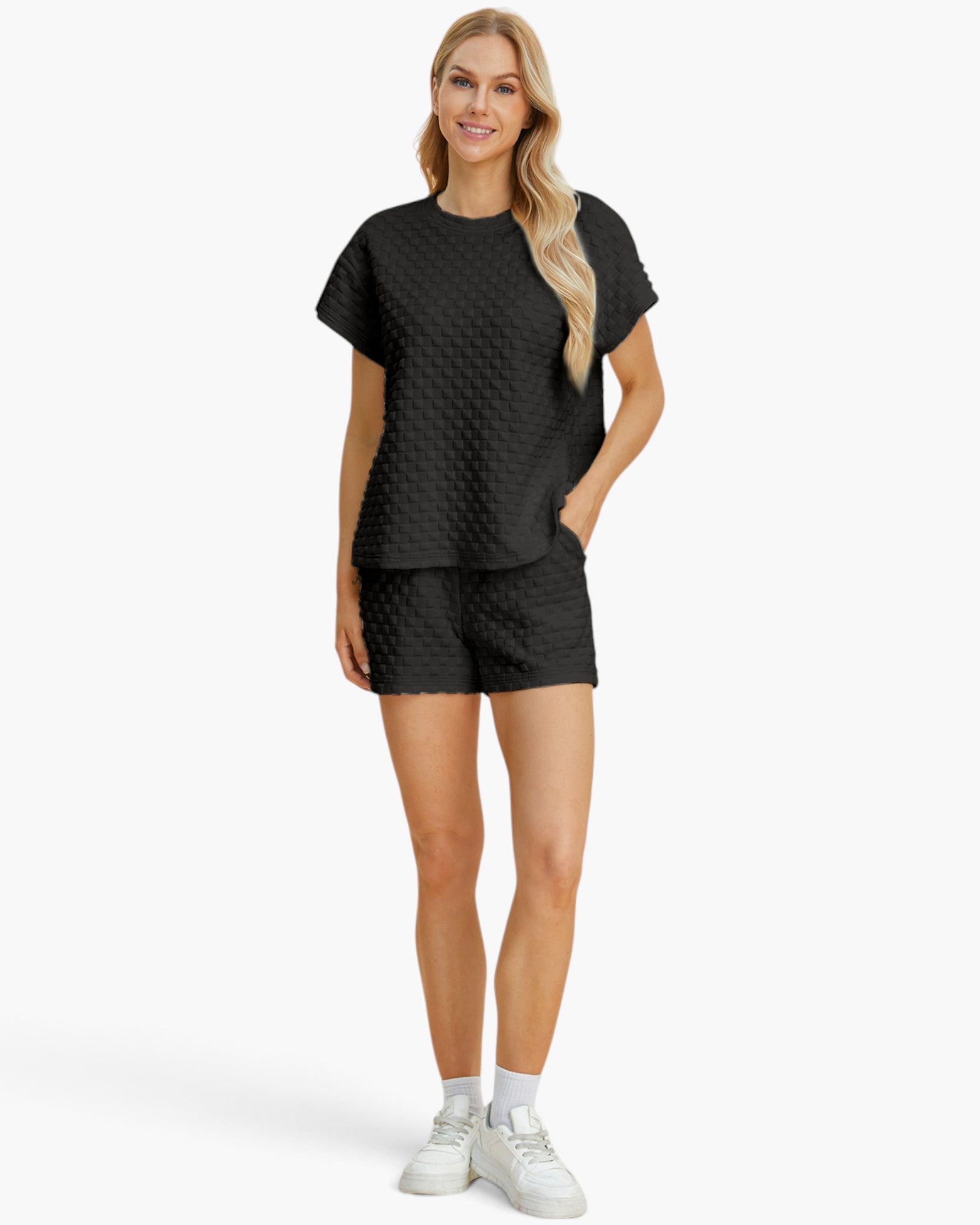 A woman modeling a matching black two-piece lounge set featuring a short-sleeve top and shorts with a textured, quilted pattern, paired with white sneakers against a light gray studio background.