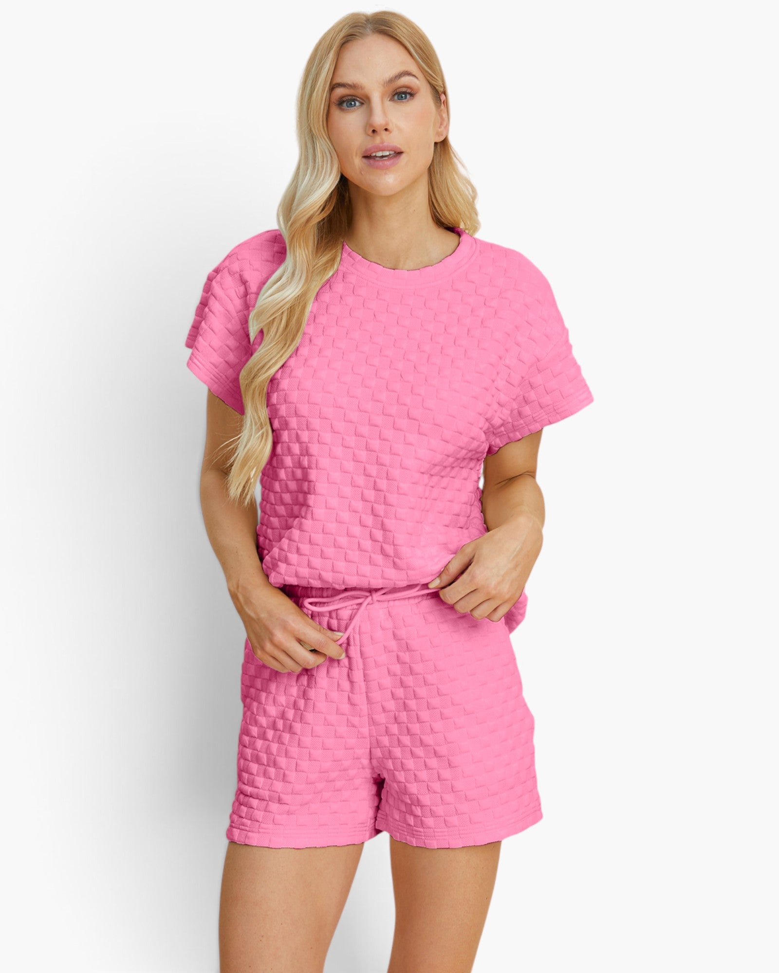 A woman modeling a bright pink two-piece lounge set with a short-sleeve top and matching shorts, both featuring a textured basketweave pattern, against a plain white studio background.