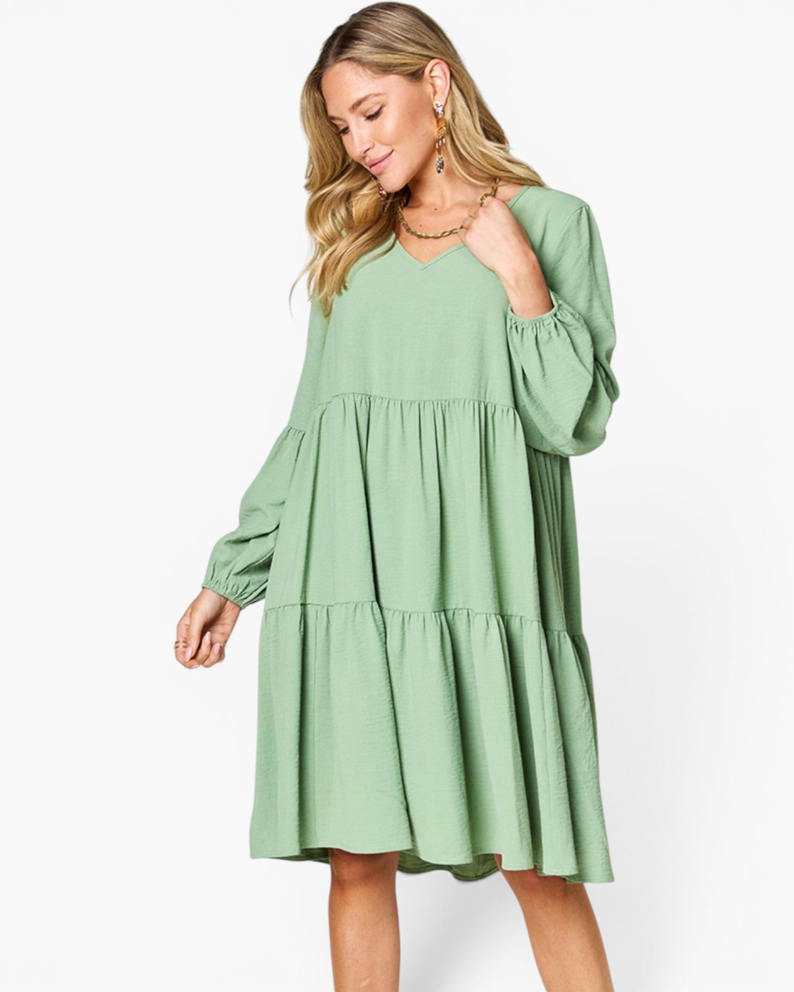A woman modeling a sage green tiered long-sleeve dress with a V-neckline, photographed against a clean white studio background.