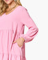 A close-up of a woman wearing a pink tiered long-sleeve dress with a V-neckline and elasticized cuffs, shown against a plain light background.