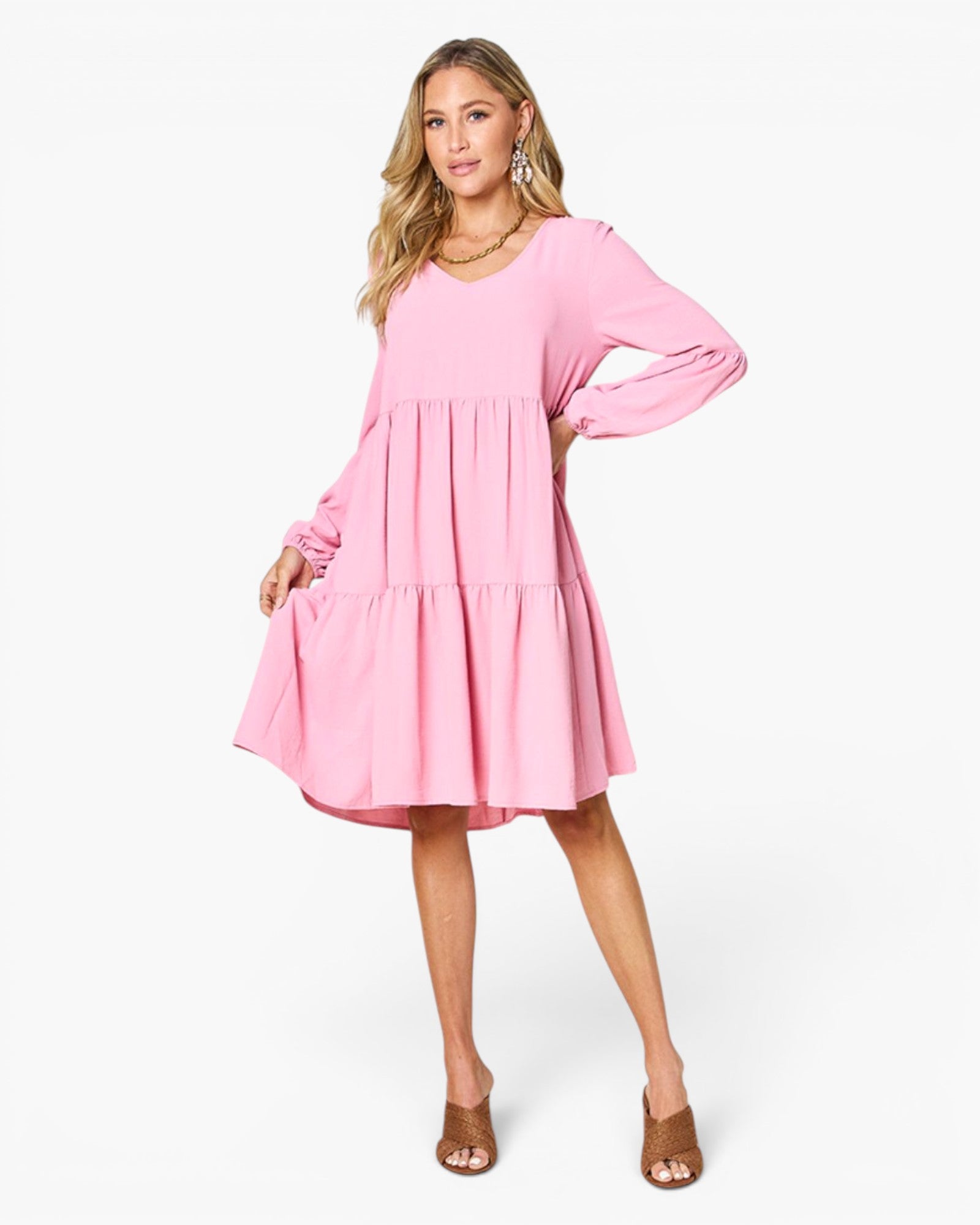 A woman modeling a light pink long-sleeve tiered dress with a scoop neckline, photographed against a clean white studio background.