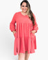A woman wearing a coral pink tiered long-sleeve dress with pockets, standing against a plain white studio background.