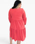 A woman viewed from the back wearing a coral pink tiered long-sleeve dress against a plain white background.