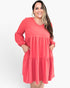 A woman wearing a coral pink tiered tunic dress with long sleeves and side pockets, standing against a plain white studio background.