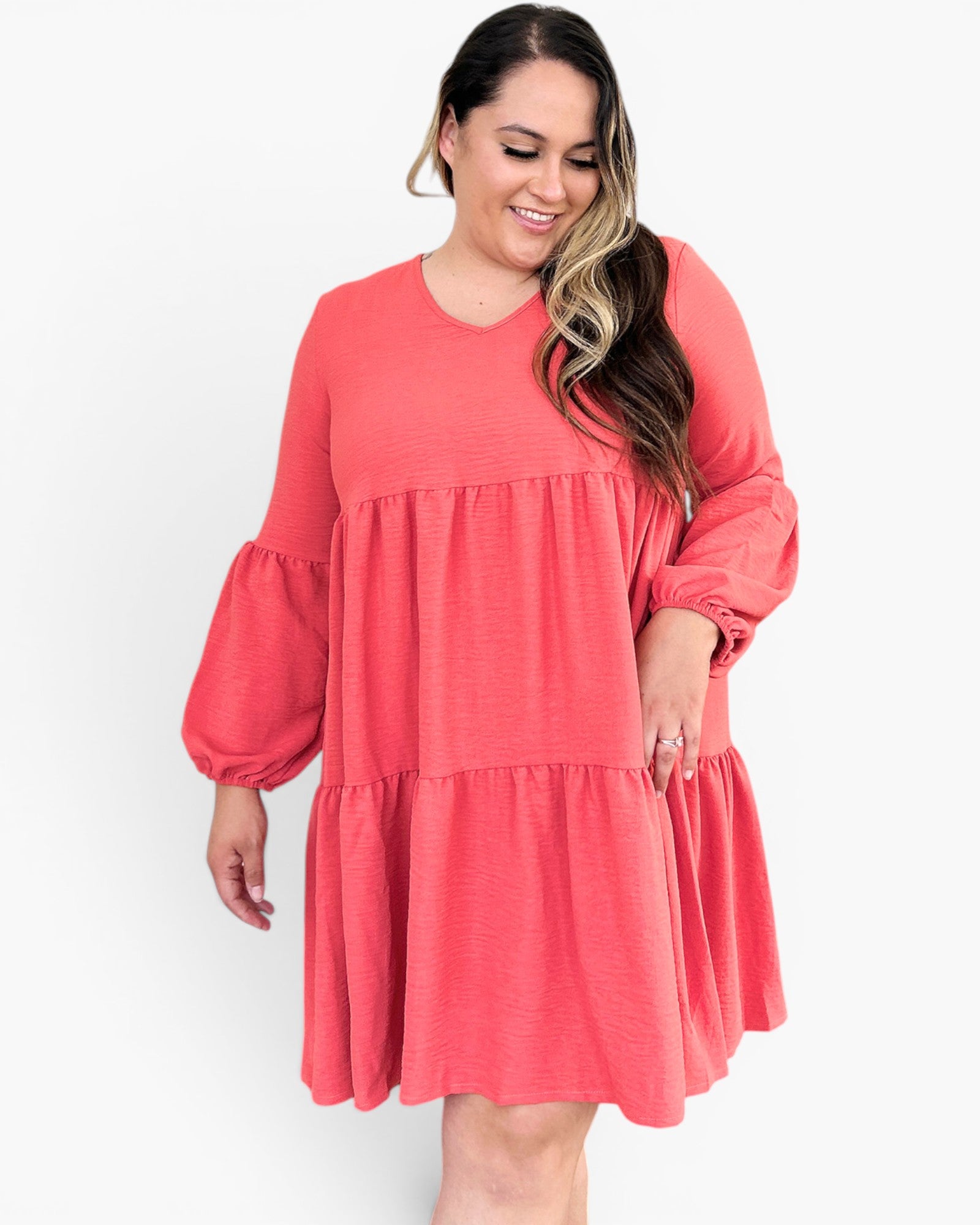 A woman wearing a coral pink tiered long-sleeve dress with balloon sleeves, shown against a plain white studio background.