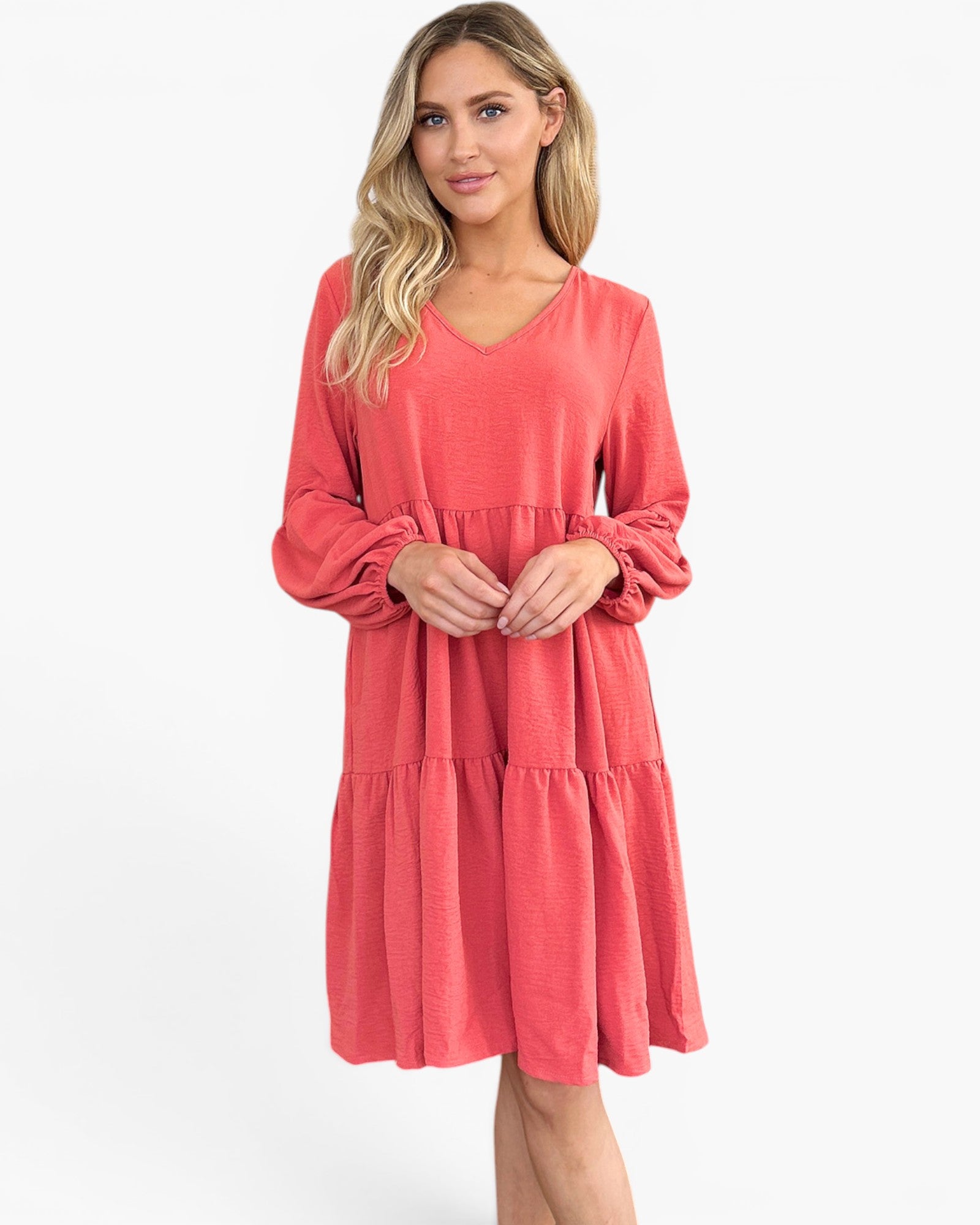 A woman modeling a coral pink tiered long-sleeve dress with a V-neckline against a plain white studio background.