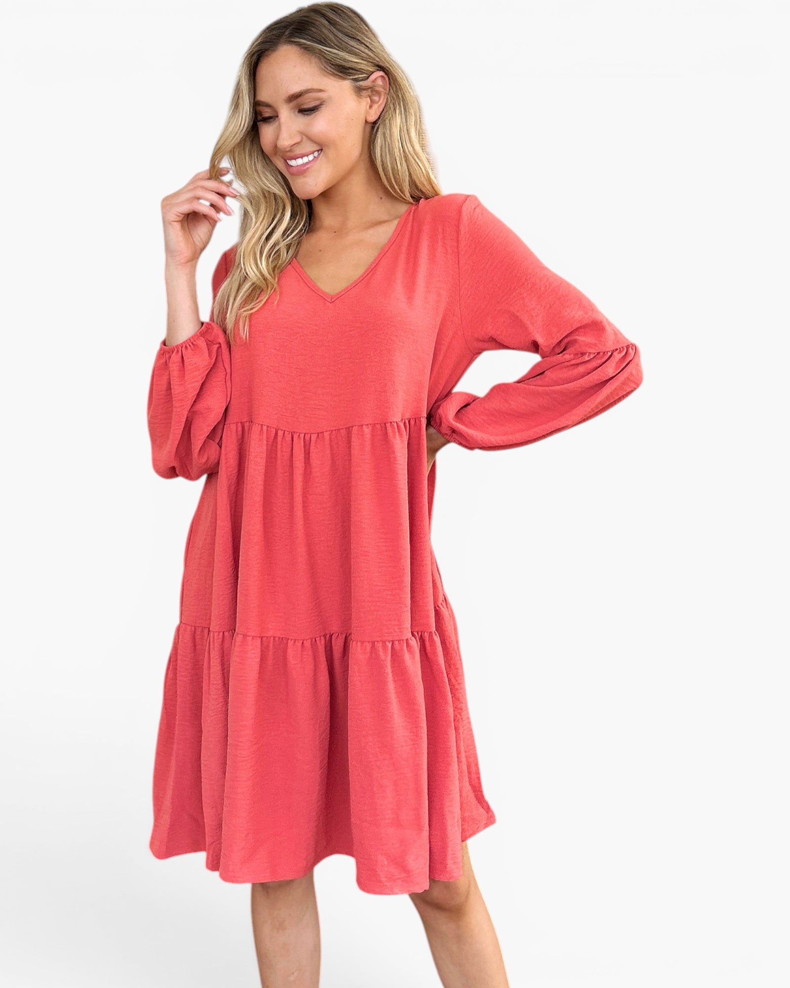 A woman modeling a coral pink tiered long-sleeve dress with a V-neckline, photographed against a plain white studio background.