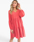 A woman modeling a coral pink tiered mini dress with long balloon sleeves and a V-neckline, set against a plain white studio background.