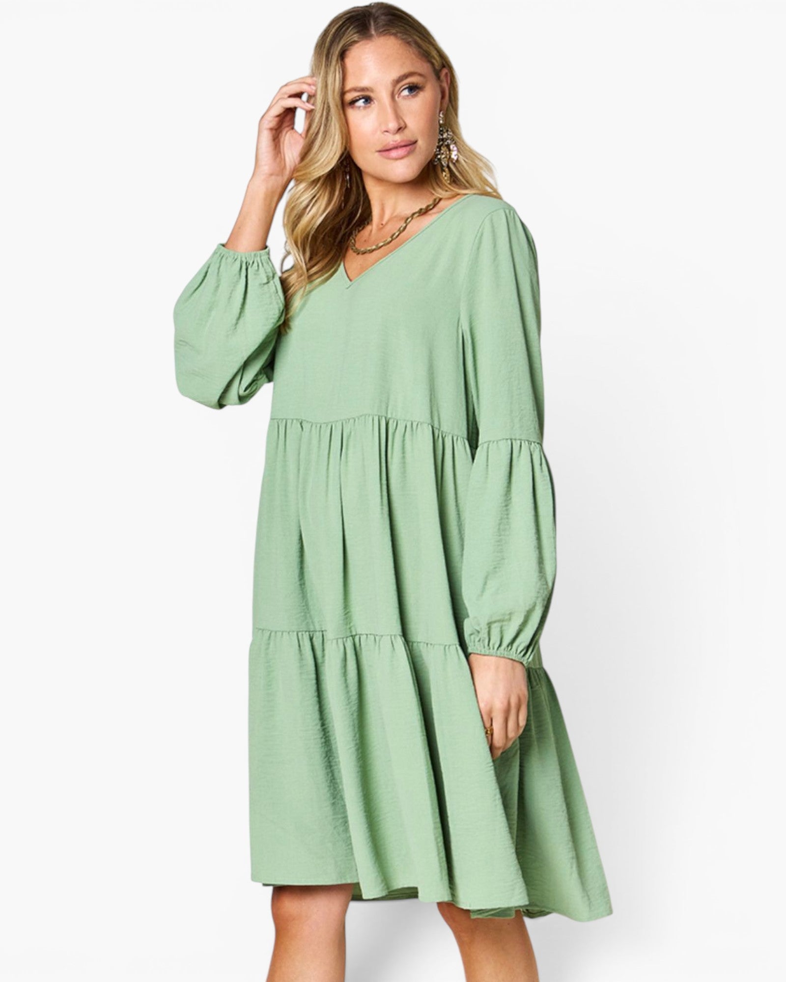 A woman modeling a light sage green tiered dress with long balloon sleeves and a V-neckline, set against a plain white studio background.