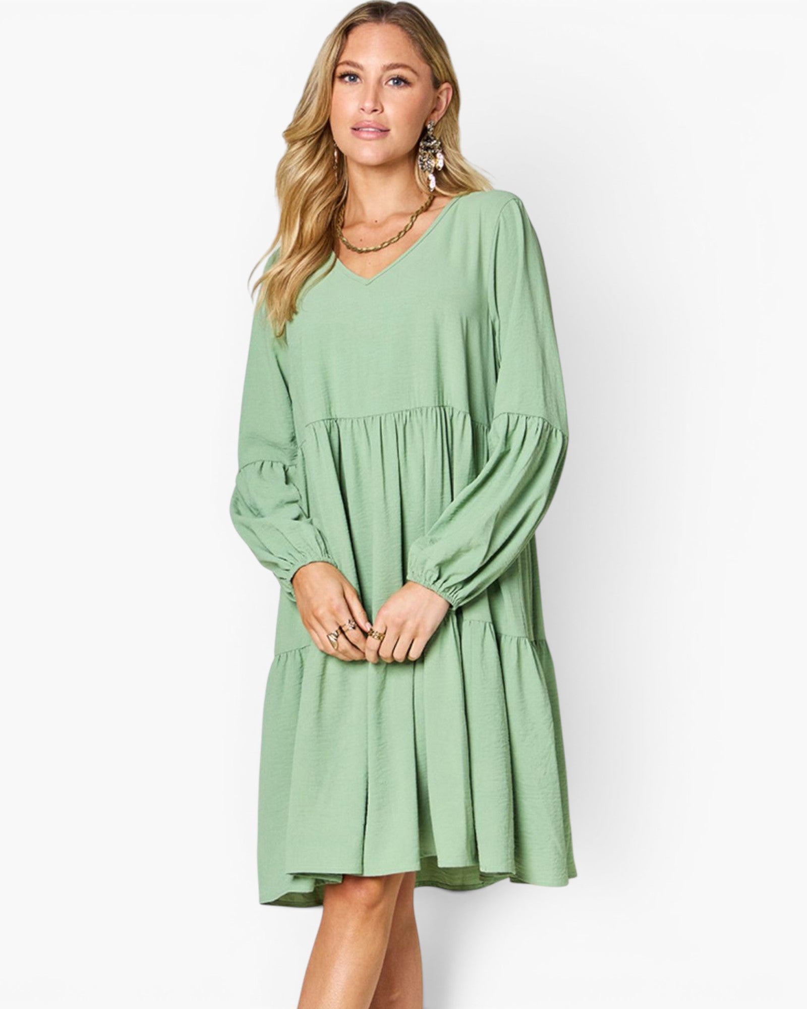 A woman modeling a sage green tiered long-sleeve dress with a V-neckline, photographed against a plain white studio background.